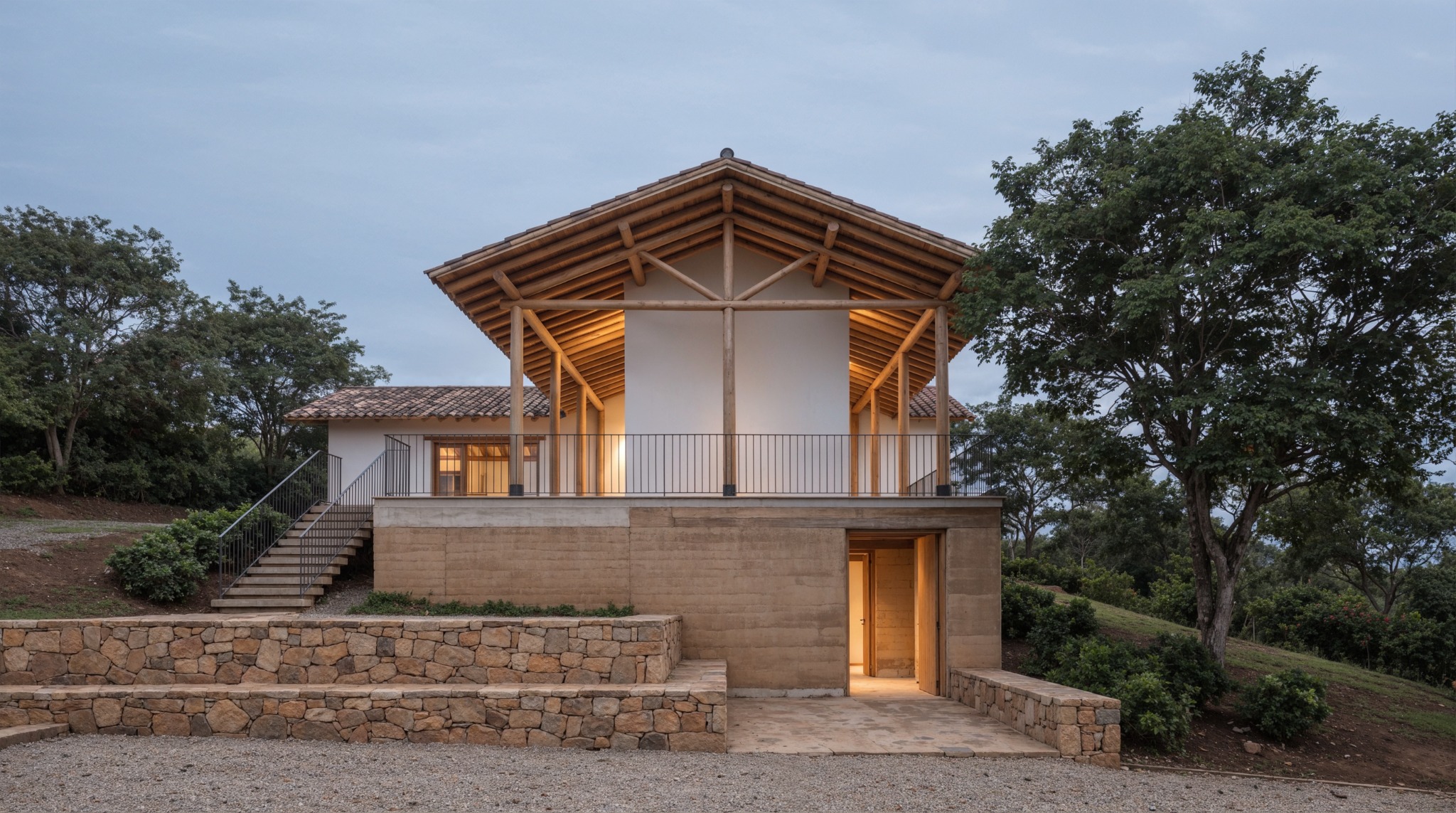 Casa rural elevada sobre basamento de tierra y piedra, con gran cubierta de madera y luz cálida al atardecer.